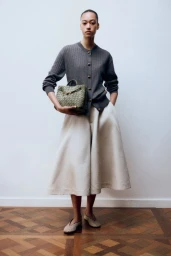 Model wearing a grey buttoned cardigan and wide cream skirt holding a woven green handbag against a plain wall