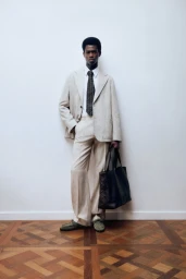 Full-length portrait of a male model wearing a light beige suit and dark patterned tie while holding a large black leather tote bag.