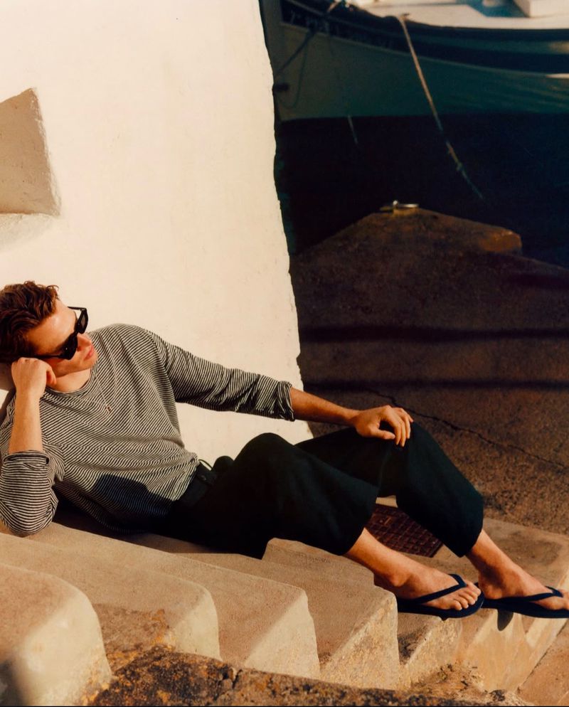 Man in sunglasses lounges on stone steps by a building near the water, wearing a striped long-sleeved shirt and dark pants with flip-flops.