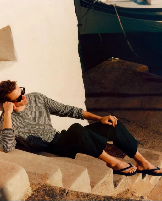 Man in sunglasses lounges on stone steps by a building near the water, wearing a striped long-sleeved shirt and dark pants with flip-flops.