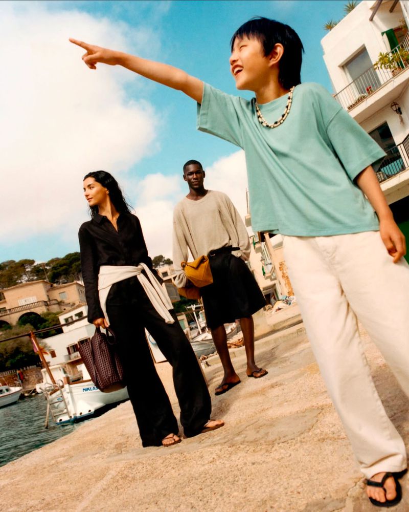 Three people stand on a stone pier near a Mediterranean town on a sunny day. An Asian child points at the sky with a smile.