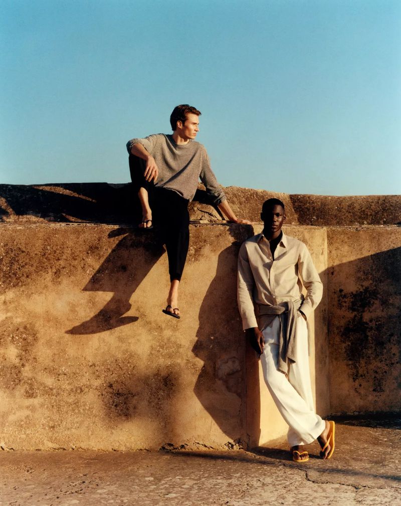 Two male models in relaxed neutral summer clothing and flip-flops posing against a sunlit textured stone wall under a clear blue sky.