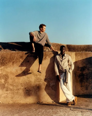 Two male models in relaxed neutral summer clothing and flip-flops posing against a sunlit textured stone wall under a clear blue sky.