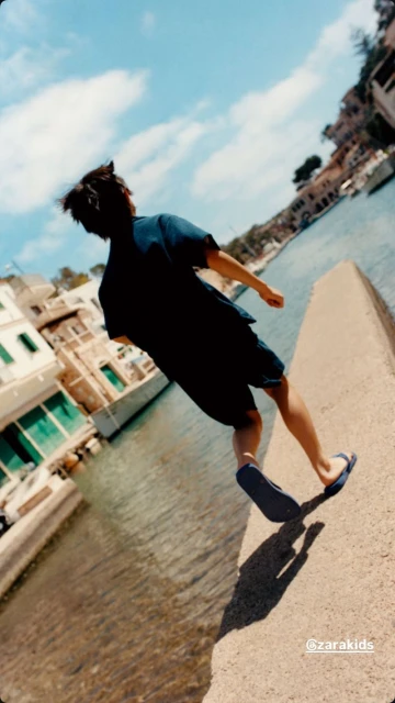A child in a blue t-shirt walks along a concrete barrier by a canal in a Mediterranean town under a blue sky with scattered clouds.