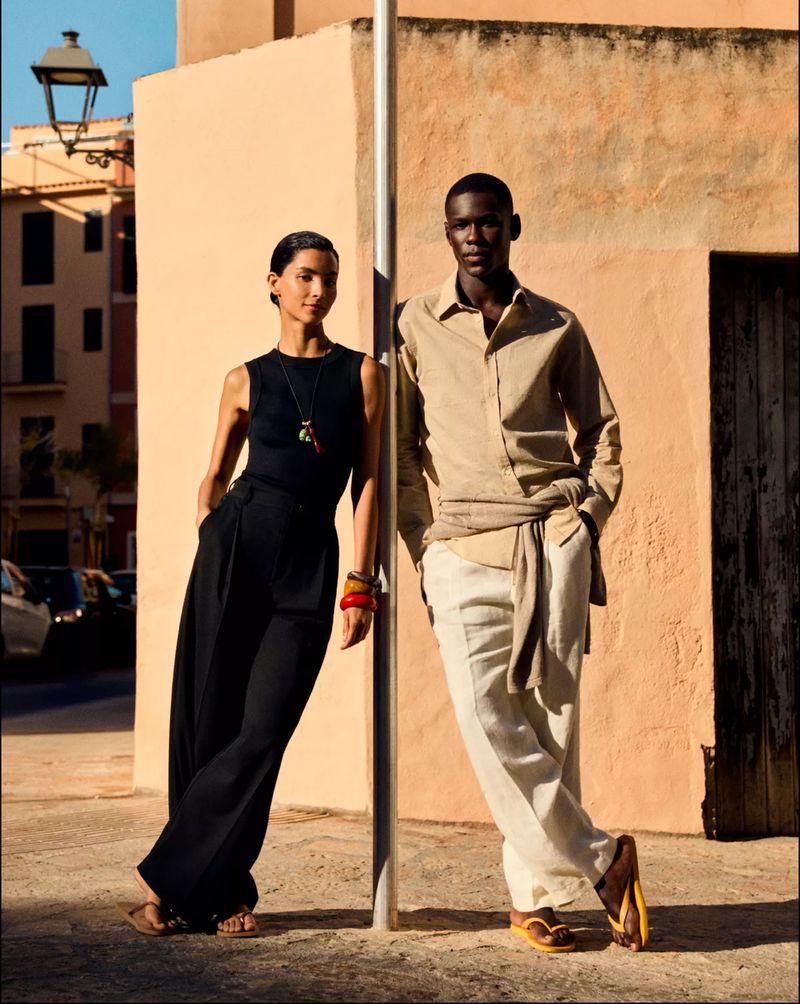 A fashionable couple poses outdoors against a peach-colored wall. The woman wears a sleeveless top and wide-leg pants; the man wears a linen shirt and trousers.