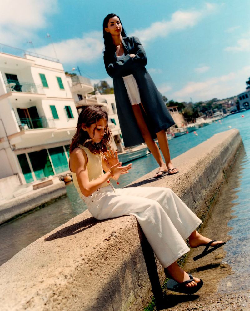 Two women in stylish casualwear pose by the waterfront with white buildings and blue sky in the background on a sunny day.