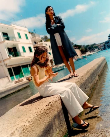 Two women in stylish casualwear pose by the waterfront with white buildings and blue sky in the background on a sunny day.