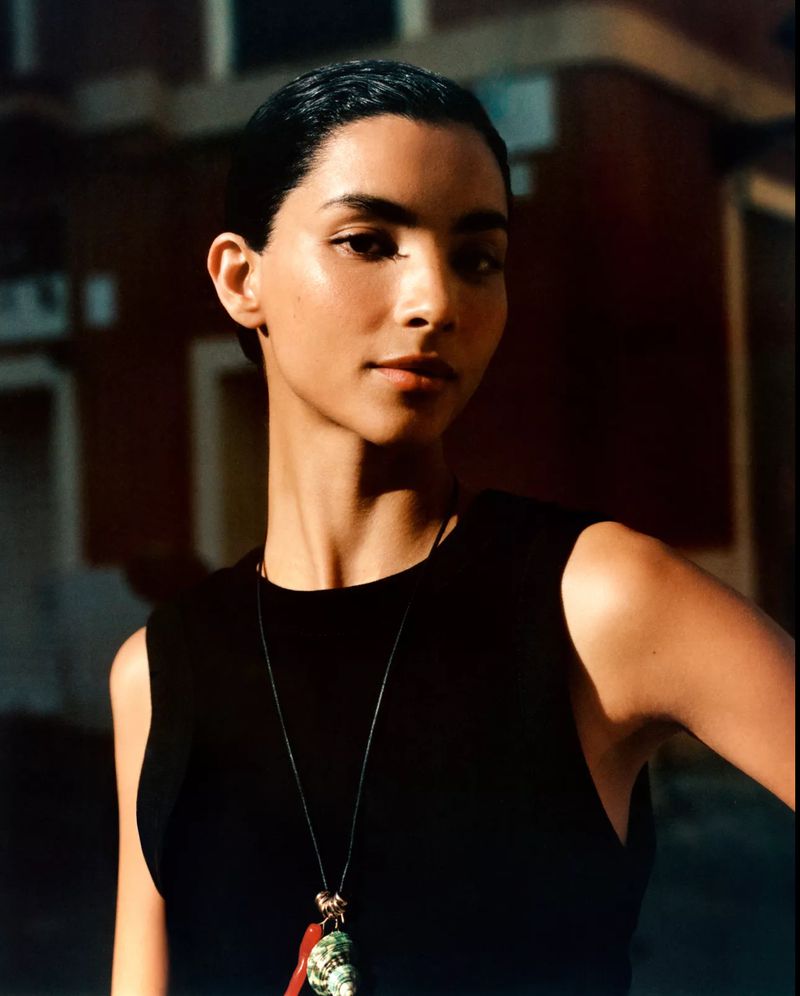 Model Tindi Mar wearing a sleeveless black top and a seashell charm necklace in warm sunlight.