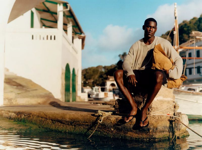 Young black man in casual clothing sitting on a concrete pier at a coastal harbor on a sunny day with boats and buildings in the background.