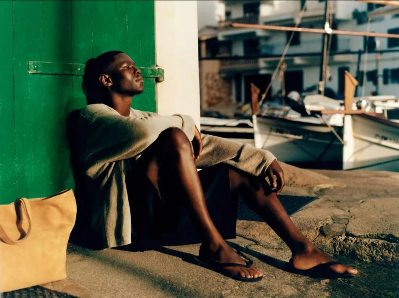 An adult male sits next to a green door wearing a sweater and sandals with a tan tote bag on a paved waterfront with boats.