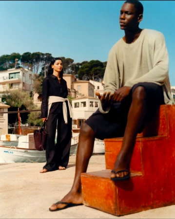Two fashion models pose outdoors near a waterfront. One sits on red steps; the other stands in black clothing with a white belt and handbag.