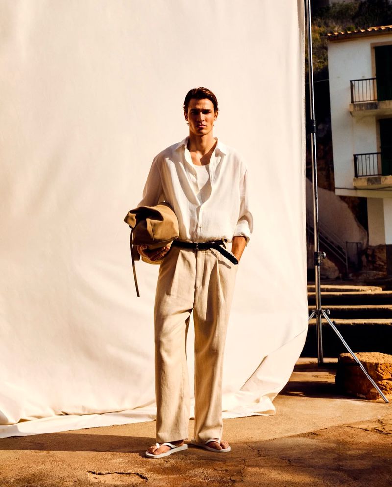A fashionable adult male standing in front of a portable backdrop, wearing a white shirt, beige pants, and carrying a neutral-toned bag.