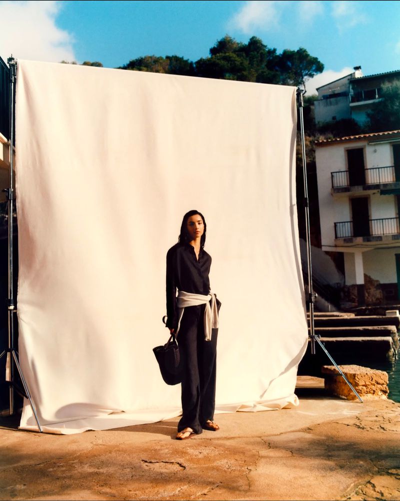 A fashionable woman in a black outfit with a sash poses on a stone surface next to a white backdrop, with buildings in the background.