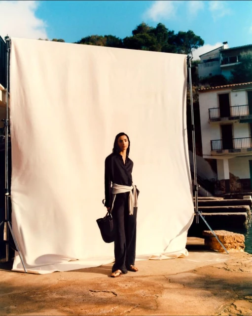 A fashionable woman in a black outfit with a sash poses on a stone surface next to a white backdrop, with buildings in the background.