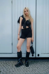 Model Stella Lucia in black crop top, shorts and tall boots holding a cylindrical bag against a white wall.