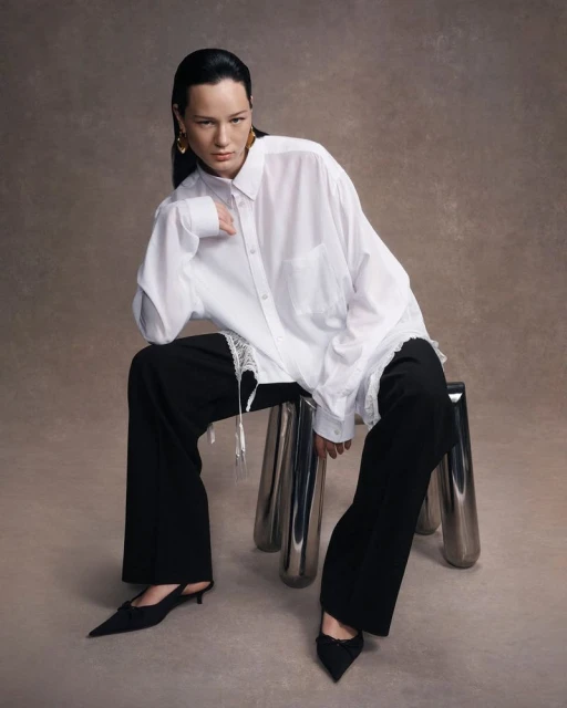 A woman sits on a chrome stool wearing a white shirt, black pants, and black shoes in a studio setting with a neutral background.