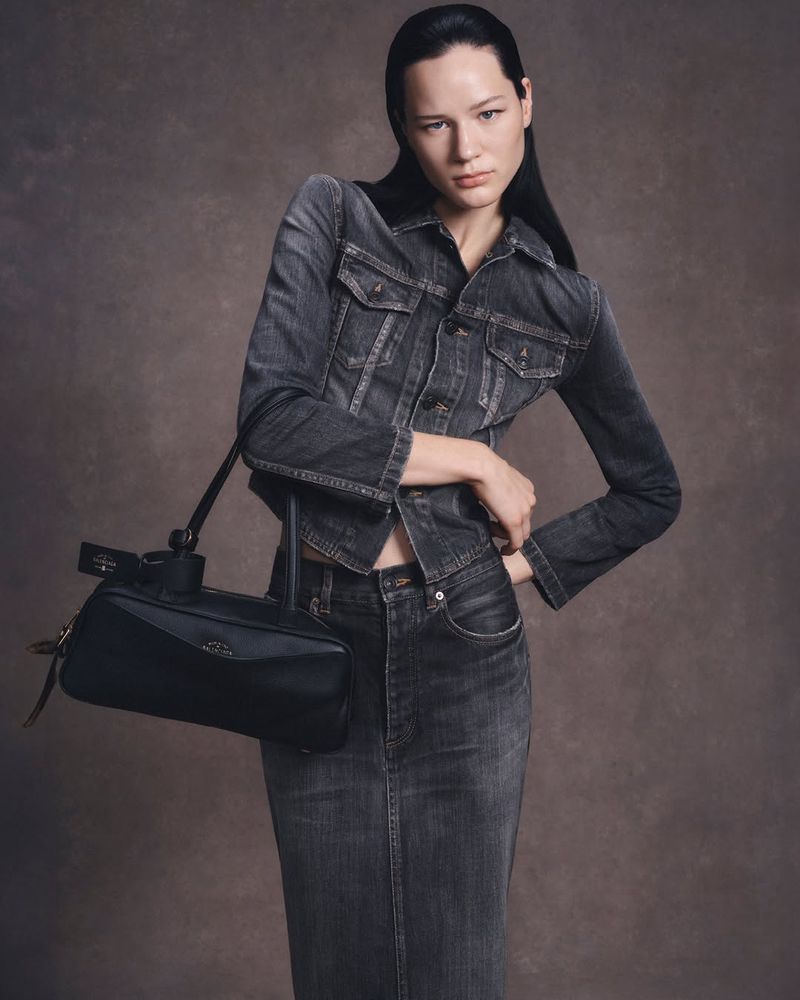 Model Lydia Celesta wearing a washed grey denim jacket and matching skirt holding a black leather bag against a neutral backdrop.