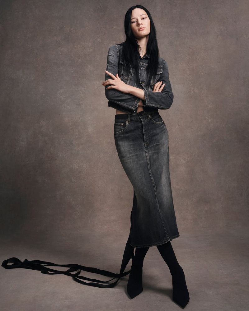 Model Lydia Celesta standing with arms crossed in a washed black denim jacket and matching long skirt against a textured taupe backdrop.