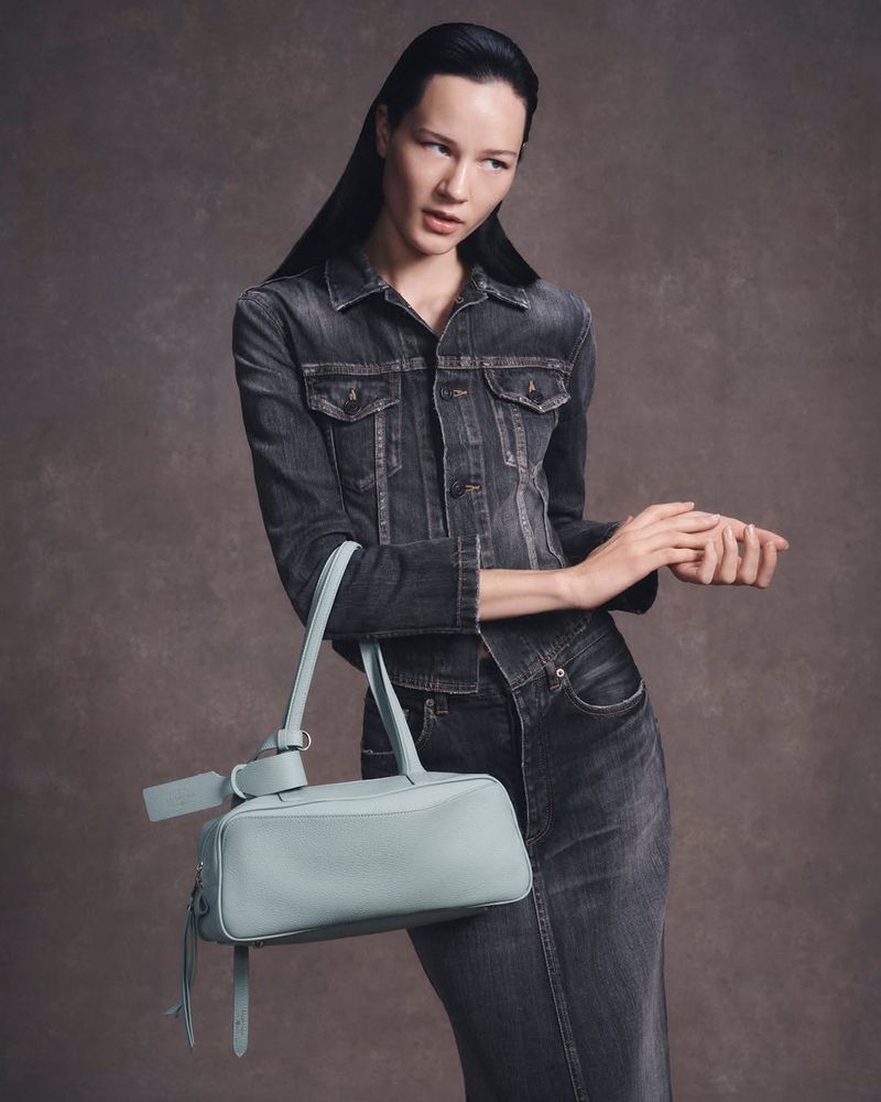 A slender fashion model poses with a light blue handbag against a neutral studio backdrop, wearing a faded black denim jacket and long skirt.