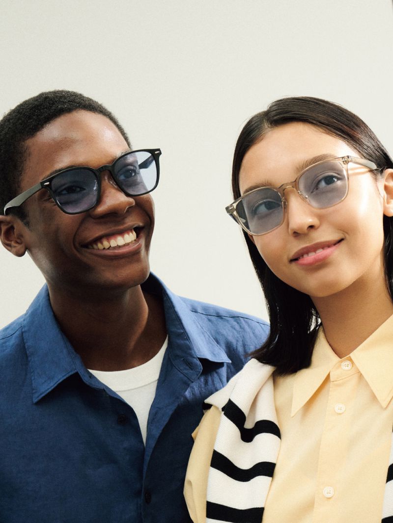 Two smiling people, one Black man and one Asian woman, wear stylish eyewear in a bright studio setting, showcasing modern fashion and inclusivity.