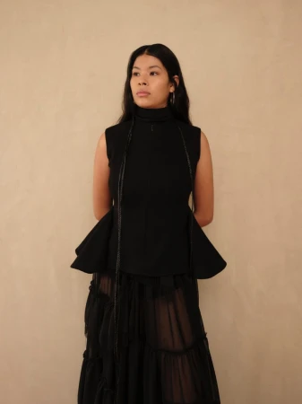 Model standing against a neutral textured background wearing a black sleeveless peplum top and layered sheer skirt