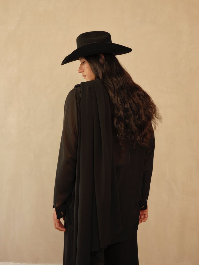 Model with long dark hair wearing a black hat and black sheer top against a neutral wall