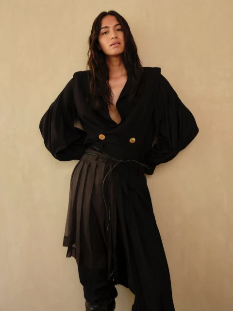 A slender, dark-haired person poses against a neutral wall, wearing a black blouse with wide sleeves and layered, draped black pants.