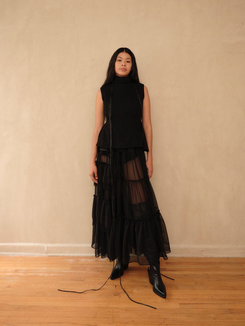 Model standing against a neutral wall wearing a black sleeveless top and sheer layered skirt
