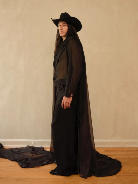Elegant person in black cowboy hat and sheer cape stands in front of neutral wall. Light brown wood floor completes simple studio shot.