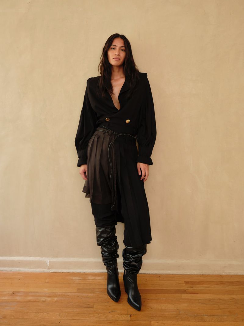 A woman poses indoors against a beige wall, wearing a black jacket with gold buttons, a layered skirt, and black knee-high scrunch boots.