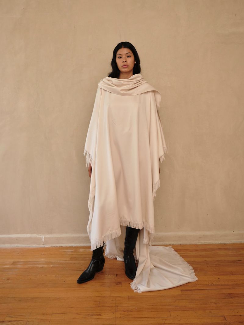 Woman poses indoors wearing draped white fabric shawl and black heeled boots against a neutral wall and wood floor backdrop.