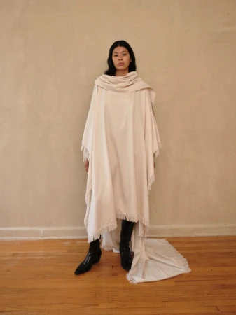 Woman poses indoors wearing draped white fabric shawl and black heeled boots against a neutral wall and wood floor backdrop.