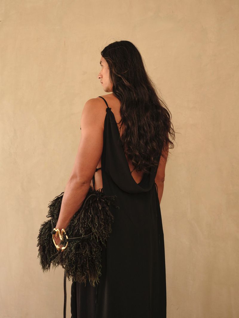 An adult in a black low-back dress, holding a feather bag, poses against a cream-colored wall in a studio setting.