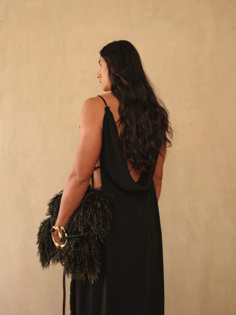 An adult in a black low-back dress, holding a feather bag, poses against a cream-colored wall in a studio setting.