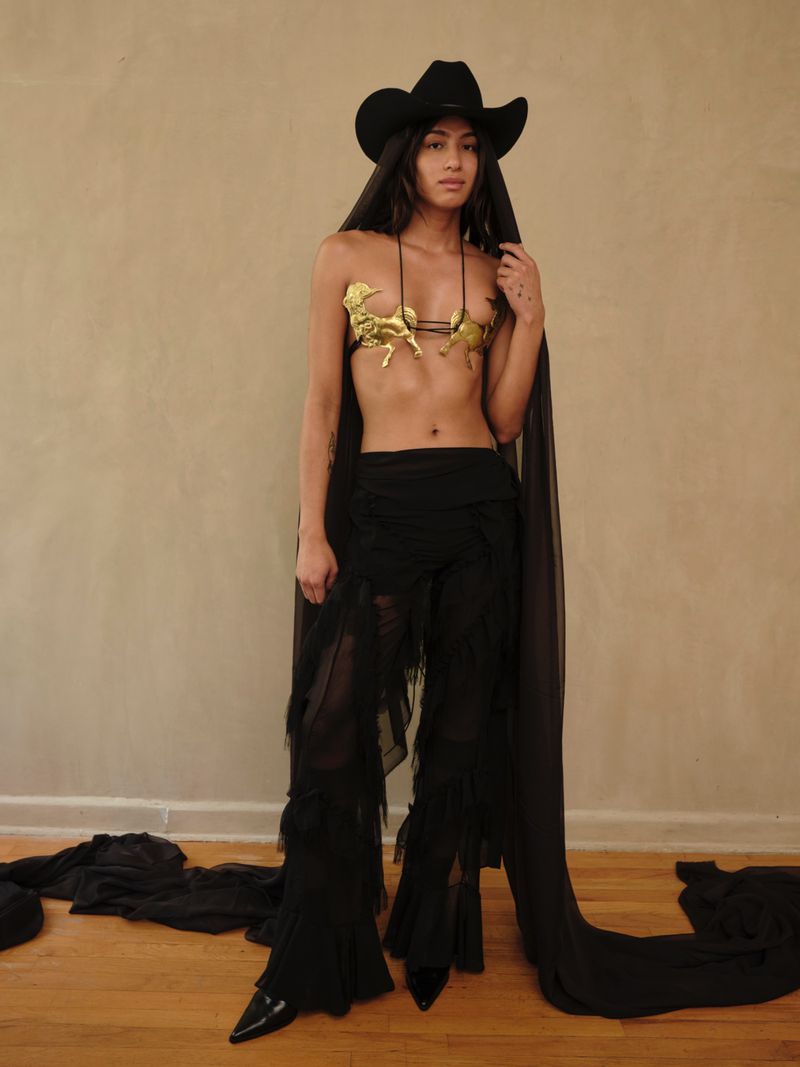 Woman poses in a gold bra with horse figures, black hat with veil, and tasseled black pants against a tan wall indoors.