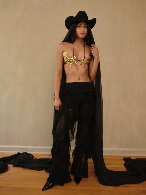 Woman poses in a gold bra with horse figures, black hat with veil, and tasseled black pants against a tan wall indoors.