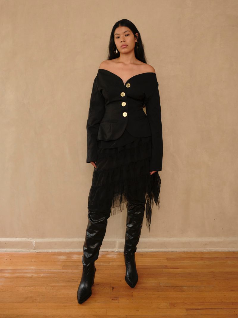 Woman poses in a black off-the-shoulder jacket with gold buttons and a layered skirt with black leather high heeled boots.