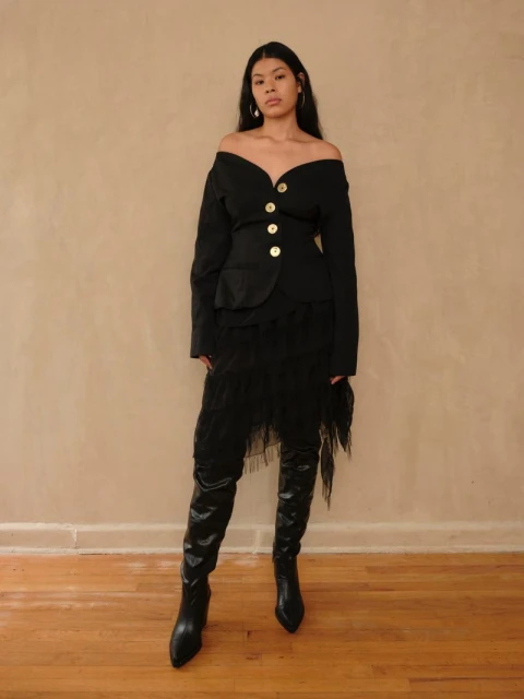 Woman poses in a black off-the-shoulder jacket with gold buttons and a layered skirt with black leather high heeled boots.