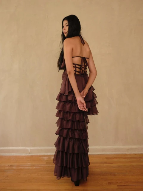 A woman wearing a brown halter top and ruffled maxi skirt stands indoors against a neutral background, looking over her shoulder.