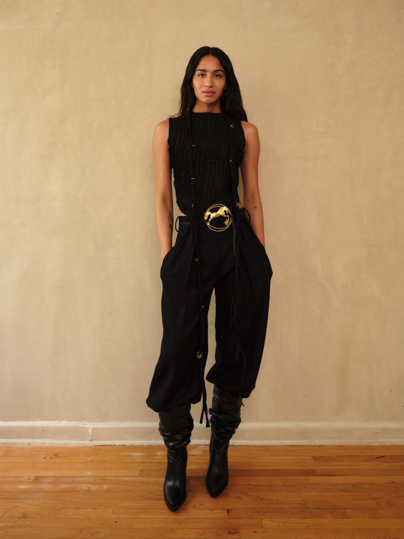Full-length portrait of a woman in a black sleeveless top, baggy black pants with a gold belt buckle, and high black leather boots.