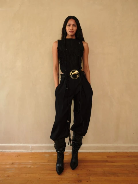 Full-length portrait of a woman in a black sleeveless top, baggy black pants with a gold belt buckle, and high black leather boots.