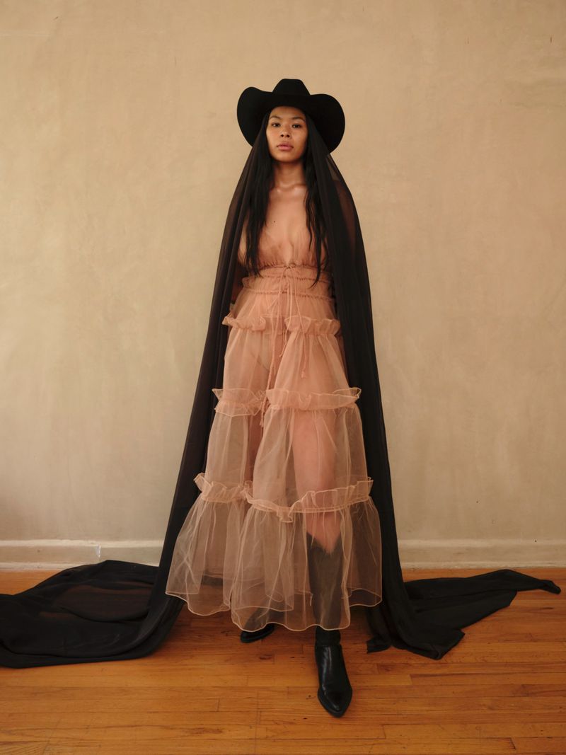 Model wearing a layered sheer ruffled dress, black cowboy hat, and long black veil cascading down to the wooden floor.