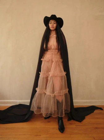 Model wearing a layered sheer ruffled dress, black cowboy hat, and long black veil cascading down to the wooden floor.