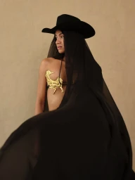 Model wearing a black cowboy hat and draped sheer black fabric with a gold sculptural chest piece against a neutral wall
