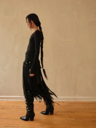 Model with a long braided hairstyle wearing a black top and skirt with tall black boots against a neutral wall and wooden floor
