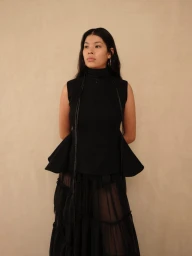 Model standing against a neutral textured background wearing a black sleeveless peplum top and layered sheer skirt