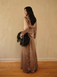 Model in a long satin dress holding a black feathered handbag against a textured wall