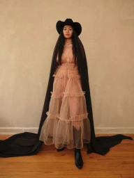 Model wearing a layered sheer ruffled dress, black cowboy hat, and long black veil cascading down to the wooden floor.