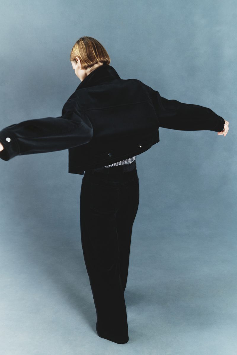 Model seen from behind with arms outstretched wearing a cropped black jacket and black trousers against a muted blue studio backdrop.
