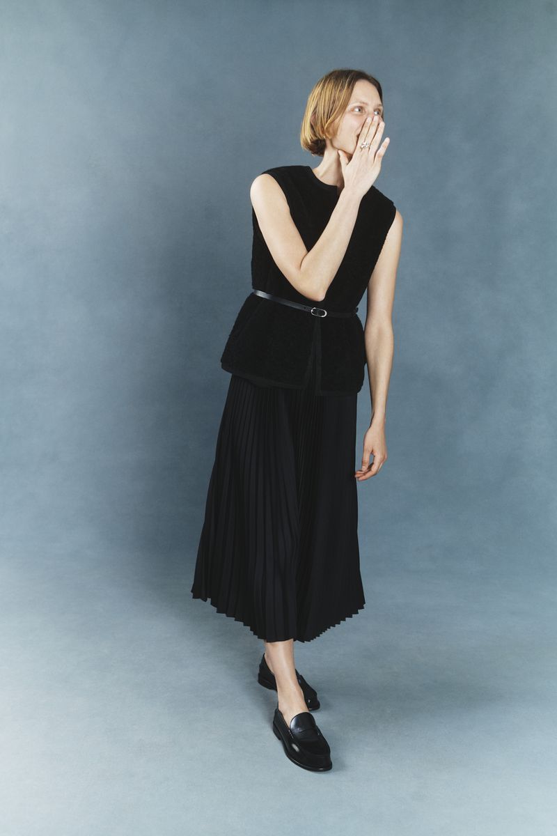An adult woman in a black sleeveless top and black pleated skirt poses against a blue backdrop, with a hand near her face.